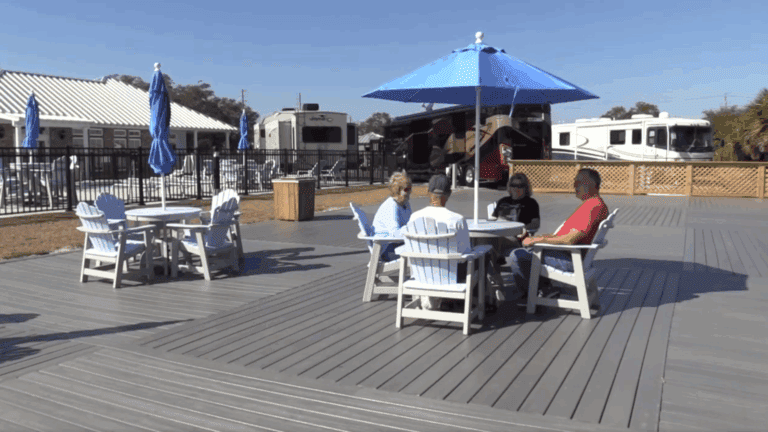 Four people sit at a round table with a blue umbrella on a deck near RVs and other empty tables, on a sunny day.