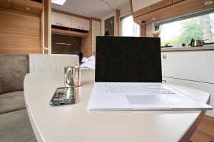 A silver laptop, smartphone, and metal mug sit on a table inside a modern camper van with wood paneling and a window. A silver laptop, smartphone, and metal mug sit on a table inside a modern camper van with wood paneling and a window.