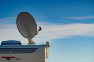 A satellite dish mounted on top of a white vehicle, likely an RV, against a clear blue sky with a few wispy clouds. A satellite dish mounted on top of a white vehicle, likely an RV, against a clear blue sky with a few wispy clouds.