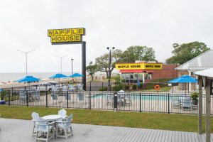 A Waffle House restaurant with a yellow sign is visible beyond a fenced outdoor pool area with lounge chairs and umbrellas, under a cloudy sky. A Waffle House restaurant with a yellow sign is visible beyond a fenced outdoor pool area with lounge chairs and umbrellas, under a cloudy sky.